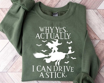 May include: An olive green sweatshirt with the text "WHY YES, ACTUALLY, I CAN DRIVE A STICK." A white graphic depicts a witch on a broom with bats and a cat. The sweatshirt has long sleeves and a crew neck.