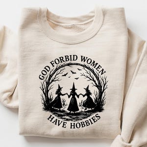May include: Sand-colored sweatshirt with a crew neck and long sleeves. The front features a black graphic of three witches holding hands within a circle of trees, with the text "GOD FORBID WOMEN HAVE HOBBIES".