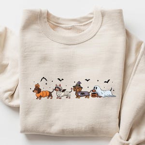 May include: A beige sweatshirt featuring a Halloween-themed dachshund design. The graphic includes four dachshunds dressed in costumes: a pumpkin, a mummy, a witch, and a ghost. Black bats and a jack-o'-lantern complete the festive design.