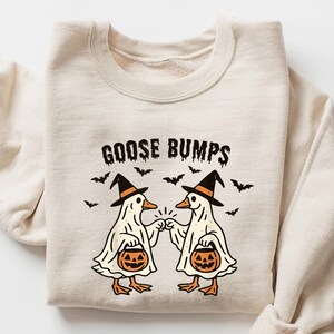 May include: Sand-colored sweatshirt with the text "GOOSE BUMPS" and a Halloween-themed graphic. The graphic features two cartoon geese dressed as ghosts, wearing witch hats, and holding jack-o'-lanterns. Small bats are scattered around the text.