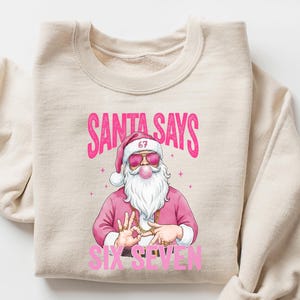 May include: Beige sweatshirt with a graphic of Santa Claus wearing sunglasses and a pink outfit. The text "SANTA SAYS SIX SEVEN" is printed in pink. The sweatshirt is folded with the sleeves rolled up.