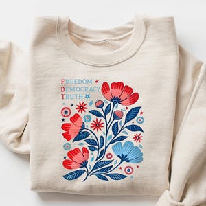 May include: A sand-colored sweatshirt with a floral design in red, blue, and white. The graphic includes the words "Freedom, Democracy, Truth". The sweatshirt has long sleeves and a crew neck.