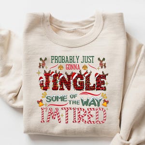 May include: A sand-colored sweatshirt with the text "Probably Just Gonna Jingle Some of the Way I'm Tired" in a festive design. The word "Jingle" is in a plaid pattern. The sweatshirt has long sleeves.