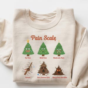 Puede incluir: Una sudadera de color arena con un gr&aacute;fico de una escala de dolor con &aacute;rboles de Navidad de dibujos animados. La escala va de "Sin dolor" a "Peor dolor posible". El texto "Pain Scale" est&aacute; arqueado sobre los &aacute;rboles.