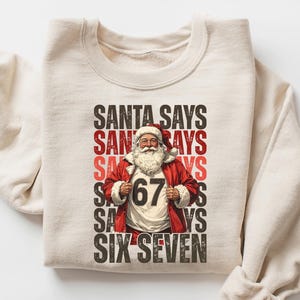 May include: A sand-colored sweatshirt with the text "SANTA SAYS" stacked vertically, with a graphic of Santa Claus wearing a red coat and the number "67" on his shirt. The sweatshirt is folded with the sleeves rolled up.