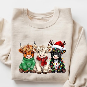 May include: A cream-colored sweatshirt featuring a graphic of three Highland cows. The cows are adorned with festive holiday attire: a green sweater, a red scarf, and a Santa hat with Christmas lights. The word "SAND" is printed in the upper right corner.