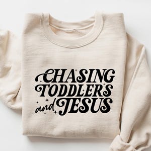 May include: A sand-colored sweatshirt with the phrase "CHASING TODDLERS and JESUS" in black script. The sweatshirt is folded with the sleeves rolled up, and the word "SAND" is in the upper right corner.