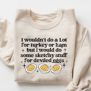 May include: Sand-colored sweatshirt with the text "I wouldn't do a Lot for turkey or ham + but I would do + some sketchy stuff for deviled eggs." The graphic includes three deviled eggs. The word "SAND" is in the upper right corner.