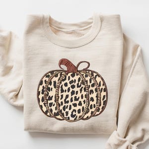 May include: Sand-colored sweatshirt featuring a pumpkin design. The pumpkin has a brown stem and a leopard print pattern in shades of brown and tan. The word "SAND" is printed in a matching color in the upper right corner.