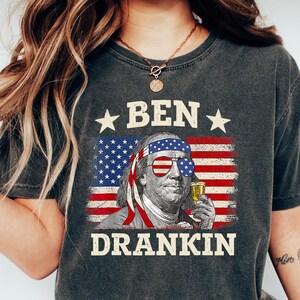 May include: Dark gray t-shirt featuring a graphic of Benjamin Franklin with sunglasses, an American flag, and the text "BEN DRANKIN". The shirt is a vintage style and is made of a soft material.