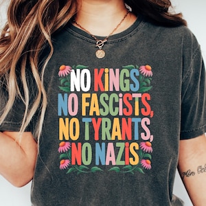 May include: Dark gray t-shirt with the text "NO KINGS, NO FASCISTS, NO TYRANTS, NO NAZIS" in colorful block letters. The text is surrounded by floral designs. The shirt is paired with blue jeans and white sneakers.
