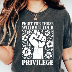 May include: Dark gray t-shirt with the text "FIGHT FOR THOSE WITHOUT YOUR PRIVILEGE" in white. The design includes a clenched fist and floral elements. The shirt is paired with blue jeans and white sneakers.