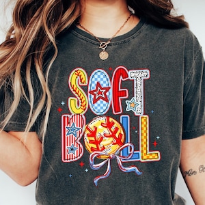Puede incluir: Una camiseta gris oscuro con un gr&aacute;fico colorido de la palabra "SOFTBALL" con una pelota de b&eacute;isbol y estrellas. La camiseta la lleva puesta una persona con el pelo largo y rubio y unos vaqueros azules.