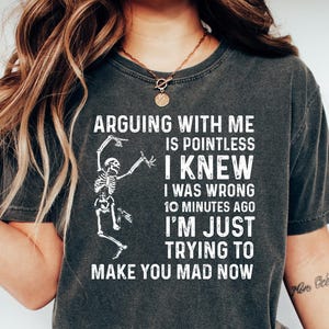 May include: Dark gray t-shirt with the text "ARGUING WITH ME IS POINTLESS I KNEW I WAS WRONG 10 MINUTES AGO I'M JUST TRYING TO MAKE YOU MAD NOW" and a skeleton graphic. The shirt is paired with blue jeans and white shoes.