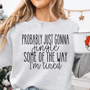 Op de afbeelding: Een lichtgrijs sweatshirt met de tekst "Probably just gonna jingle some of the way I'm tired" in het zwart. Een witte mok met een kerstman-afbeelding wordt in de hand gehouden. Een kerstboom staat op de achtergrond.