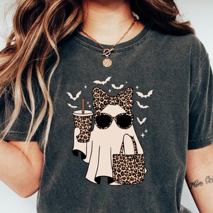 May include: Dark gray t-shirt featuring a ghost character wearing sunglasses and a leopard print bow, holding a coffee cup and a tote bag. The design includes bats and is a fun Halloween theme.
