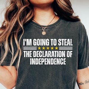May include: Dark gray t-shirt with the text "I'M GOING TO STEAL THE DECLARATION OF INDEPENDENCE" in white. The shirt has a crew neck and short sleeves. The text is above a line of five gold stars.