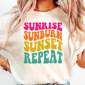 May include: Cream-colored t-shirt with the words "SUNRISE SUNBURN SUNSET REPEAT" in a retro font. The text is stacked vertically in bright pink, orange, yellow, and teal. The shirt is paired with light blue denim jeans.