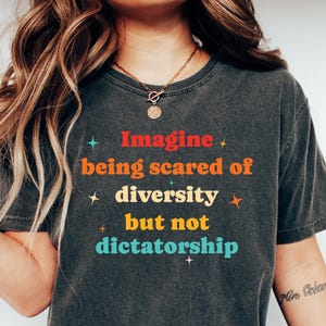 May include: A person wearing a dark grey t-shirt with the text "Imagine being scared of diversity but not dictatorship" printed on it in colorful letters. The shirt has a distressed look and is paired with blue jeans.