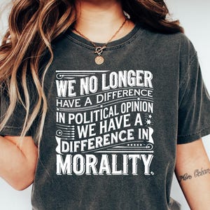 May include: Dark gray t-shirt with the text "WE NO LONGER HAVE A DIFFERENCE IN POLITICAL OPINION WE HAVE A DIFFERENCE IN MORALITY" in white. The shirt is paired with blue jeans and white sneakers.