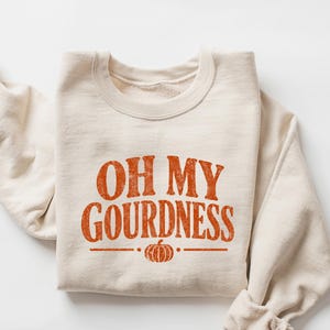 May include: A sand-colored crewneck sweatshirt featuring the orange text OH MY GOURDNESS printed in a distressed font across the chest. Below the text is a small orange pumpkin graphic centered between two horizontal lines. The word SAND is visible.