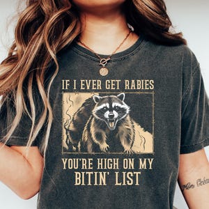 May include: Dark gray t-shirt featuring a raccoon illustration with the text "IF I EVER GET RABIES YOU'RE HIGH ON MY BITIN' LIST". The raccoon is depicted with an open mouth and sharp teeth. The shirt is worn with blue jeans and white shoes.