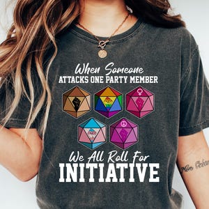 May include: Dark gray t-shirt with the text "When Someone Attacks One Party Member, We All Roll For Initiative." The shirt features colorful dice graphics representing various symbols. The person is wearing blue jeans and white sneakers.