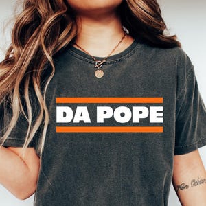 May include: Dark gray t-shirt with the text "DA POPE" in white letters, framed by two orange lines. The shirt is worn with blue jeans and white slip-on shoes. The overall style is casual and relaxed.