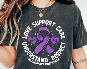 Może przedstawiać: Szary t-shirt z nadrukiem fioletowej wstążki i tekstem "Love Support Care Understand Respect Alzheimer's Awareness" w kółku wokół wstążki. Wstążka ma dwa serca, jedno z napisem "End", a drugie z napisem "ALZ".