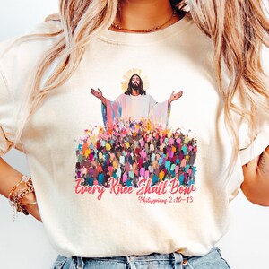 May include: A white t-shirt with a colorful graphic of Jesus Christ with the text "Every Knee Shall Bow" and "Philippians 2:10-13" printed on the front.