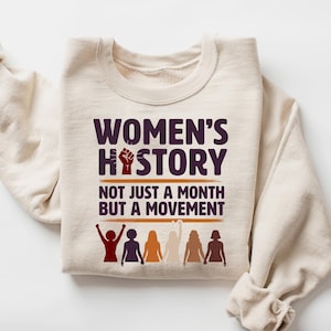 May include: A sand-colored sweatshirt with the text "WOMEN'S HISTORY NOT JUST A MONTH BUT A MOVEMENT" in dark purple and red. The design includes a raised fist and silhouettes of women in various colors.