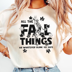 May include: Cream-colored t-shirt with the text "ALL THE FALL THINGS OR WHATEVER BLINK 182 SAYS" in black. The design includes a skeleton and maple leaves. The person is wearing light blue jeans.
