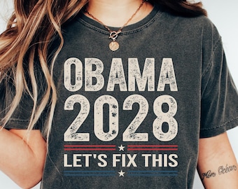 May include: A dark gray t-shirt with the text "Obama 2028" and "Let's Fix This" in white with red and blue accents. The shirt has a distressed look.