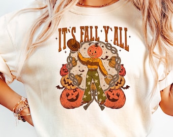 May include: Cream-colored t-shirt with the text "IT'S FALL Y'ALL" in brown lettering. The graphic features a pumpkin-headed figure holding a hat, surrounded by pumpkins and pretzels. The figure is wearing a brown shirt and green pants.