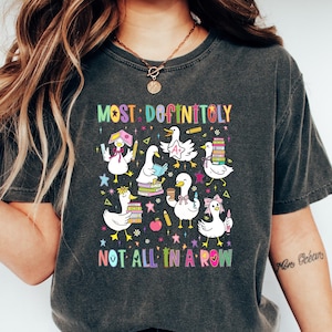 May include: Dark gray t-shirt with a colorful graphic of cartoon ducks with books, pencils, and bows. The text on the shirt reads "MOST DEFINITOLY NOT ALL IN A ROW". The shirt is worn with blue jeans and white shoes.