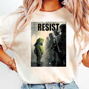 May include: Cream-colored t-shirt featuring a graphic of a frog wearing a blue scarf, facing a line of riot police. The word "RESIST" is printed above the frog. The shirt is paired with light blue jeans.