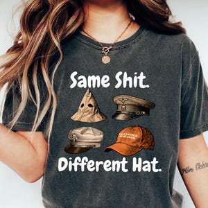 May include: Dark gray t-shirt with the text "Same Shit. Different Hat." and illustrations of four different hats. The hats include a KKK hood, a military cap, a sailor hat, and a baseball cap.