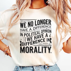 May include: Cream-colored t-shirt with the text "WE NO LONGER HAVE A DIFFERENCE IN POLITICAL OPINION WE HAVE A DIFFERENCE IN MORALITY" in black. The shirt is paired with light blue denim jeans.