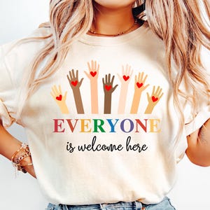 May include: Cream-colored t-shirt with the text "EVERYONE is welcome here" in rainbow colors. The shirt features a graphic of hands in various skin tones, each with a red heart.