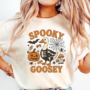 May include: Cream-colored t-shirt with a Halloween-themed graphic. The design features a goose with a skeleton body, a jack-o'-lantern, spiderwebs, and the words "Spooky Goosey" in orange. The shirt is paired with light blue jeans.
