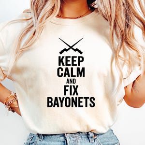 May include: Cream-colored t-shirt with the text "KEEP CALM AND FIX BAYONETS" in black. Above the text is a graphic of two crossed bayonets. The shirt is paired with light blue denim jeans.