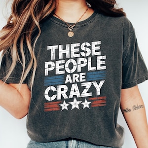 May include: Dark gray t-shirt with the text "THESE PEOPLE ARE CRAZY" in white block letters, with blue and red stripes and three white stars. The shirt is paired with blue jeans and white sneakers.
