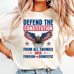 May include: Cream-colored t-shirt featuring a patriotic design. The graphic includes an eagle with outstretched wings, the text "DEFEND THE CONSTITUTION" in blue and red, and the phrase "FROM ALL ENEMIES BOTH FOREIGN AND DOMESTIC".