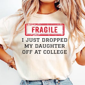 May include: Cream-colored t-shirt with the text "FRAGILE" in a red rectangle, and the phrase "I JUST DROPPED MY DAUGHTER OFF AT COLLEGE" in black. The person is wearing light blue jeans.