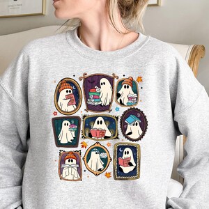May include: A light gray sweatshirt featuring a colorful graphic of cartoon ghosts reading books. Each ghost is framed in a different ornate border. Some ghosts wear hats. The design has a fall theme with leaves.