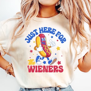 May include: Cream-colored t-shirt with the text "JUST HERE FOR the WIENERS" and a cartoon hot dog character. The hot dog has a red bun, yellow hot dog, and is giving a thumbs up. Blue jeans.