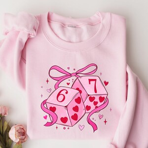 May include: A light pink sweatshirt featuring a graphic of two dice with the numbers 6 and 7. The dice are pink with red heart details and are tied together with a pink ribbon. The sweatshirt is a solid light pink color.