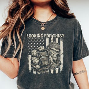 May include: Dark gray t-shirt with a graphic featuring a Bigfoot-like creature holding a 10mm object in front of an American flag design. The text "LOOKING FOR THIS?" is above the graphic. The shirt is worn by a person.
