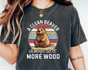 May include: A woman wearing a dark grey t-shirt with a vintage-style graphic of a beaver and the text "A Clean Beaver Always Gets More Wood".