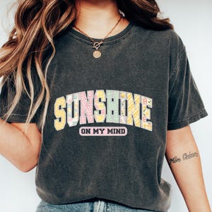 May include: Dark gray t-shirt with the text "SUNSHINE ON MY MIND" in colorful, patterned letters. The shirt is paired with blue jeans and white sneakers, creating a casual, everyday look.